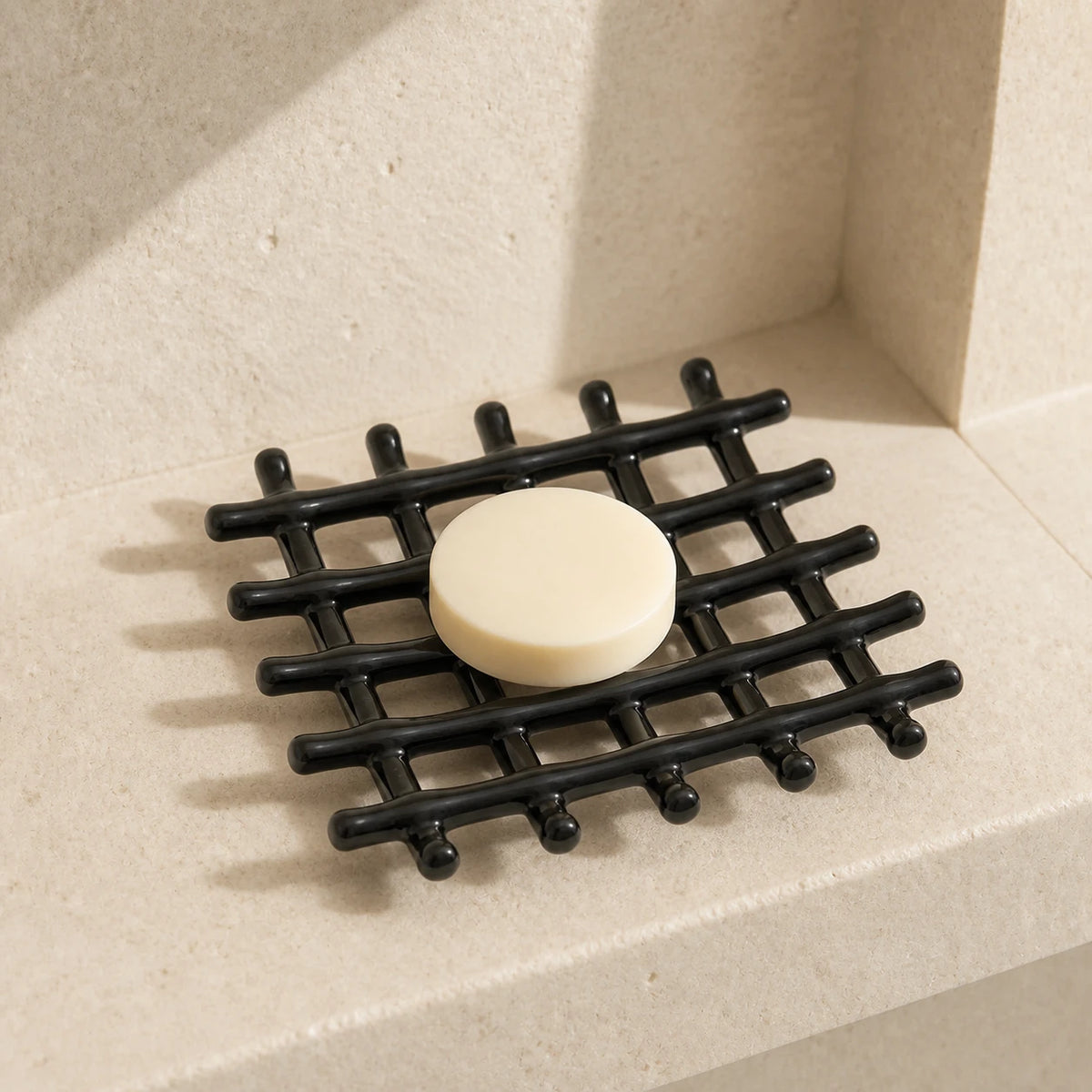 Porte-savon en treillis noir brillant avec savon rond posé sur un rebord en calcaire ensoleillé